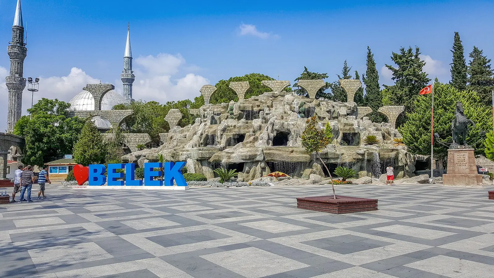 Belek town square with the BELEK sign, a rock fountain and a minaret under a blue sky